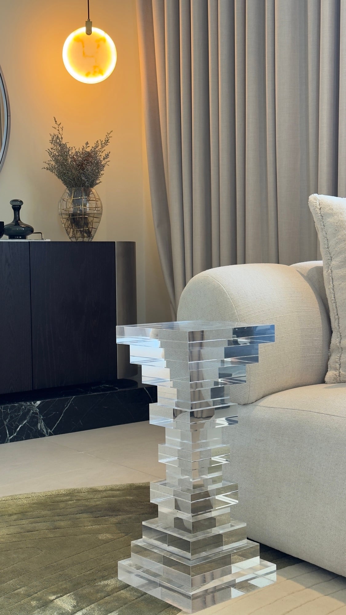 A stacked clear acrylic side table beside a cream sofa, with a glowing amber globe pendant and a dark console behind, designed by Radwa Interiors.