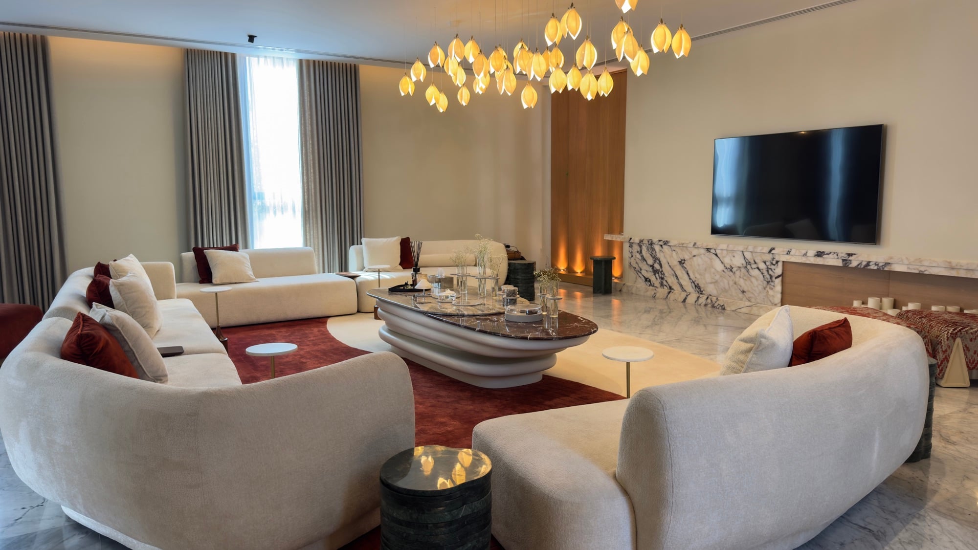 A living room with curved cream sofas around an oval stone table, under a wide chandelier of amber glass leaves, beside a marble media wall, designed by Radwa Interiors.