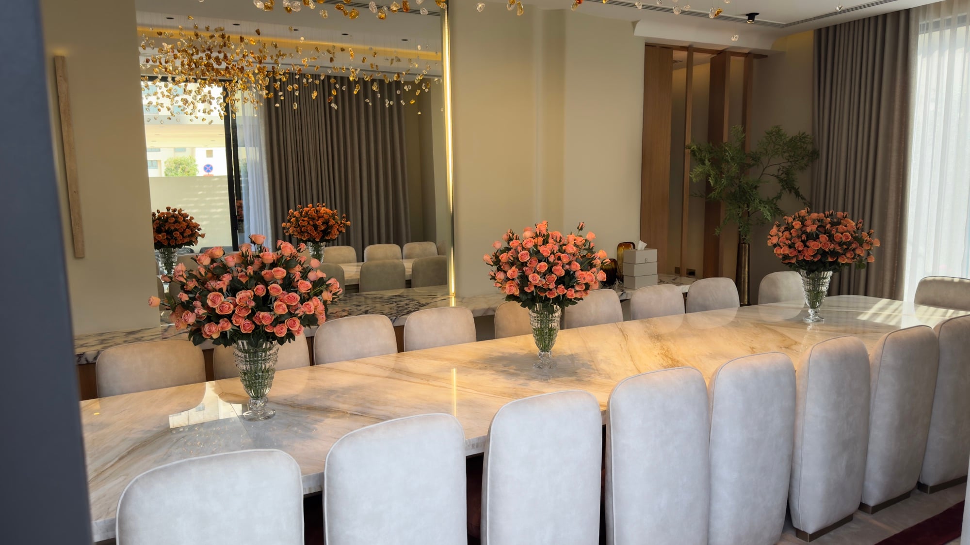 A long marble dining table lined with cream chairs and set with coral rose arrangements, reflected in a full-height mirrored wall, designed by Radwa Interiors.