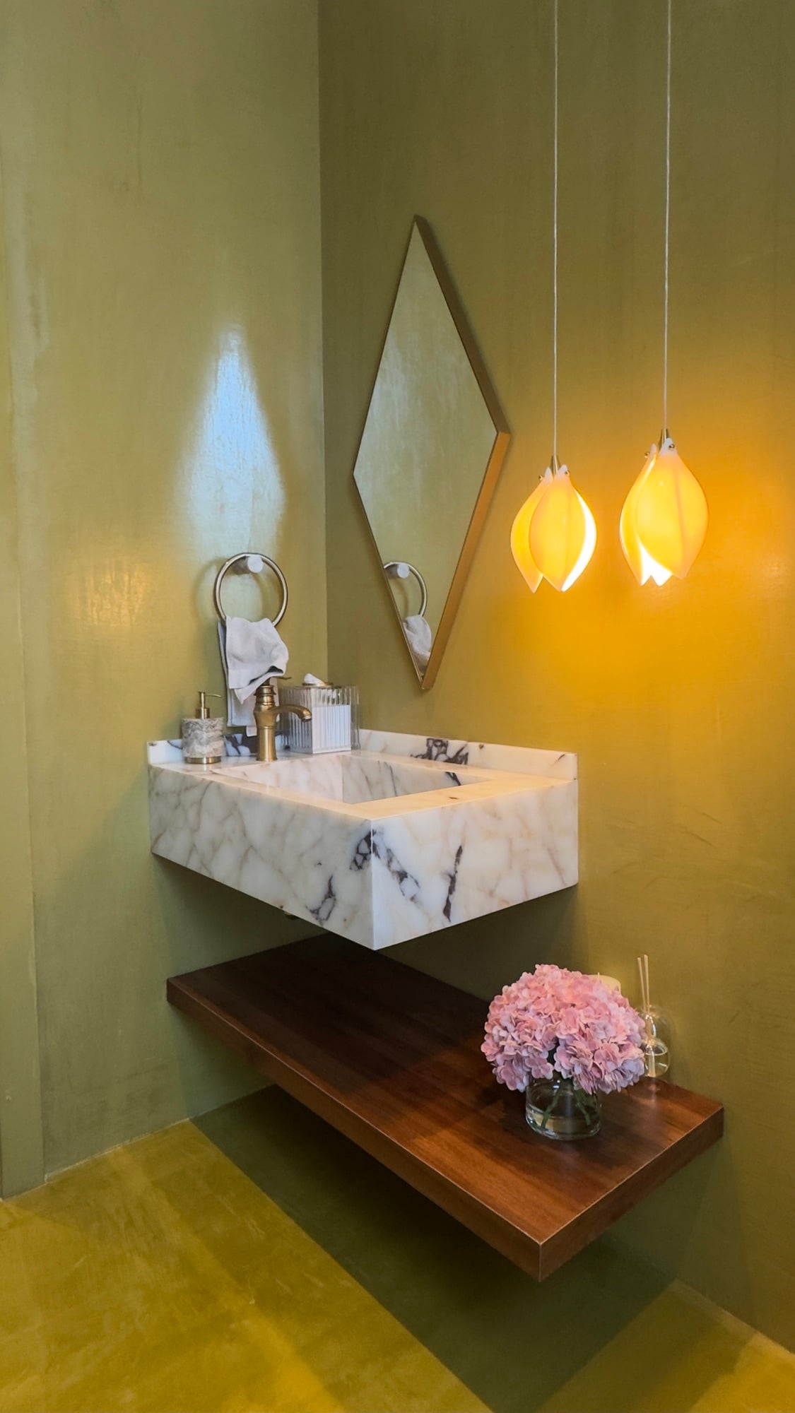A cloakroom with a marble basin on an olive-green wall, a diamond mirror and two amber petal pendants above a walnut shelf, designed by Radwa Interiors.