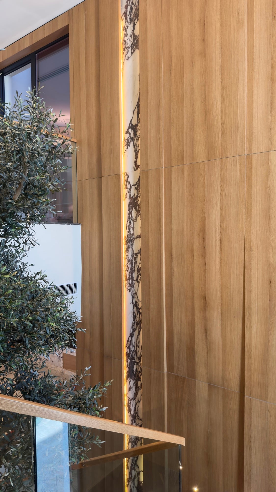 A slender illuminated onyx strip set into a timber-panelled double-height wall, beside an olive tree and a glass balustrade, designed by Radwa Interiors.