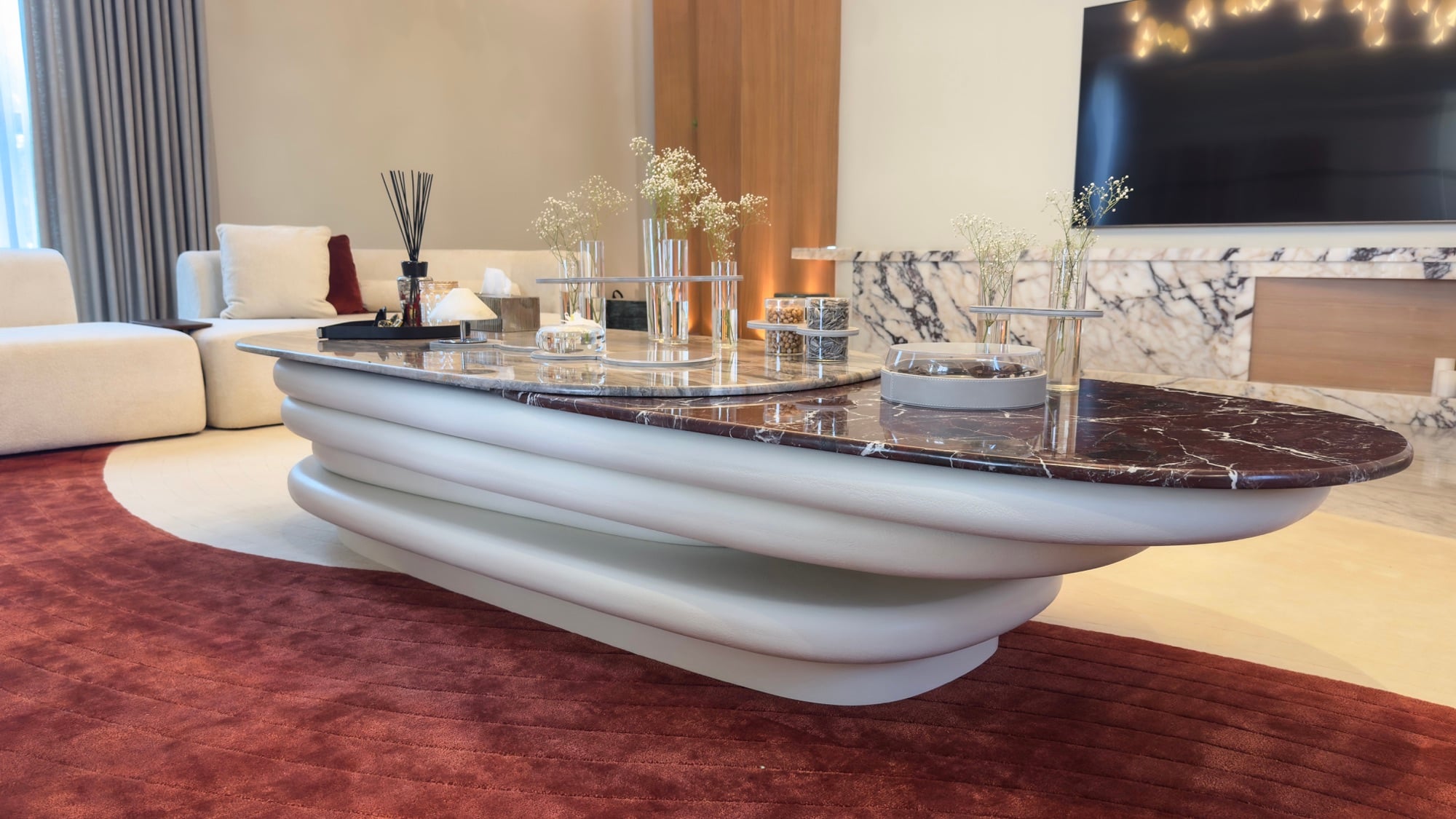 A sculptural oval coffee table with a burgundy marble top on a deep red rug, in front of a marble media wall, designed by Radwa Interiors.