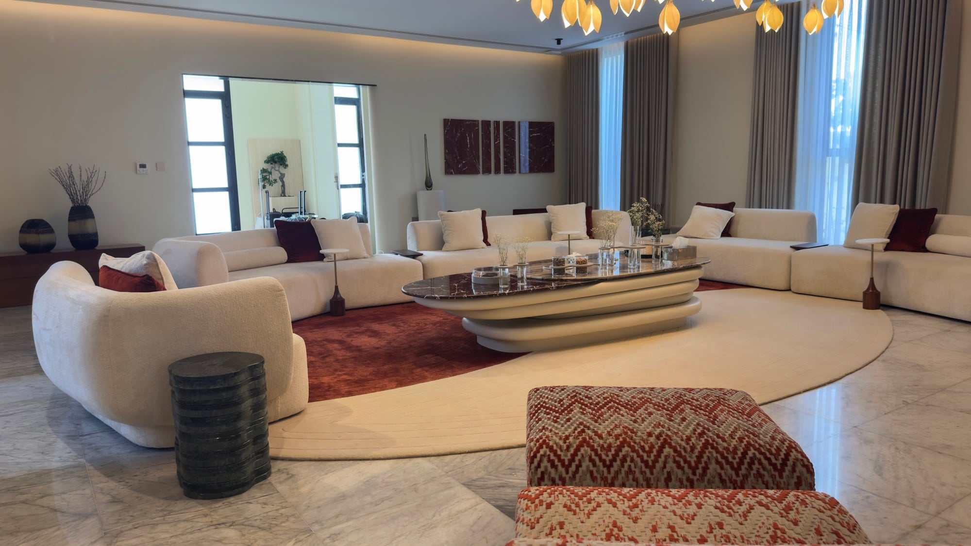 A wide living room with cream sofas on layered red and ivory rugs, patterned ottomans and an amber leaf chandelier overhead, designed by Radwa Interiors.