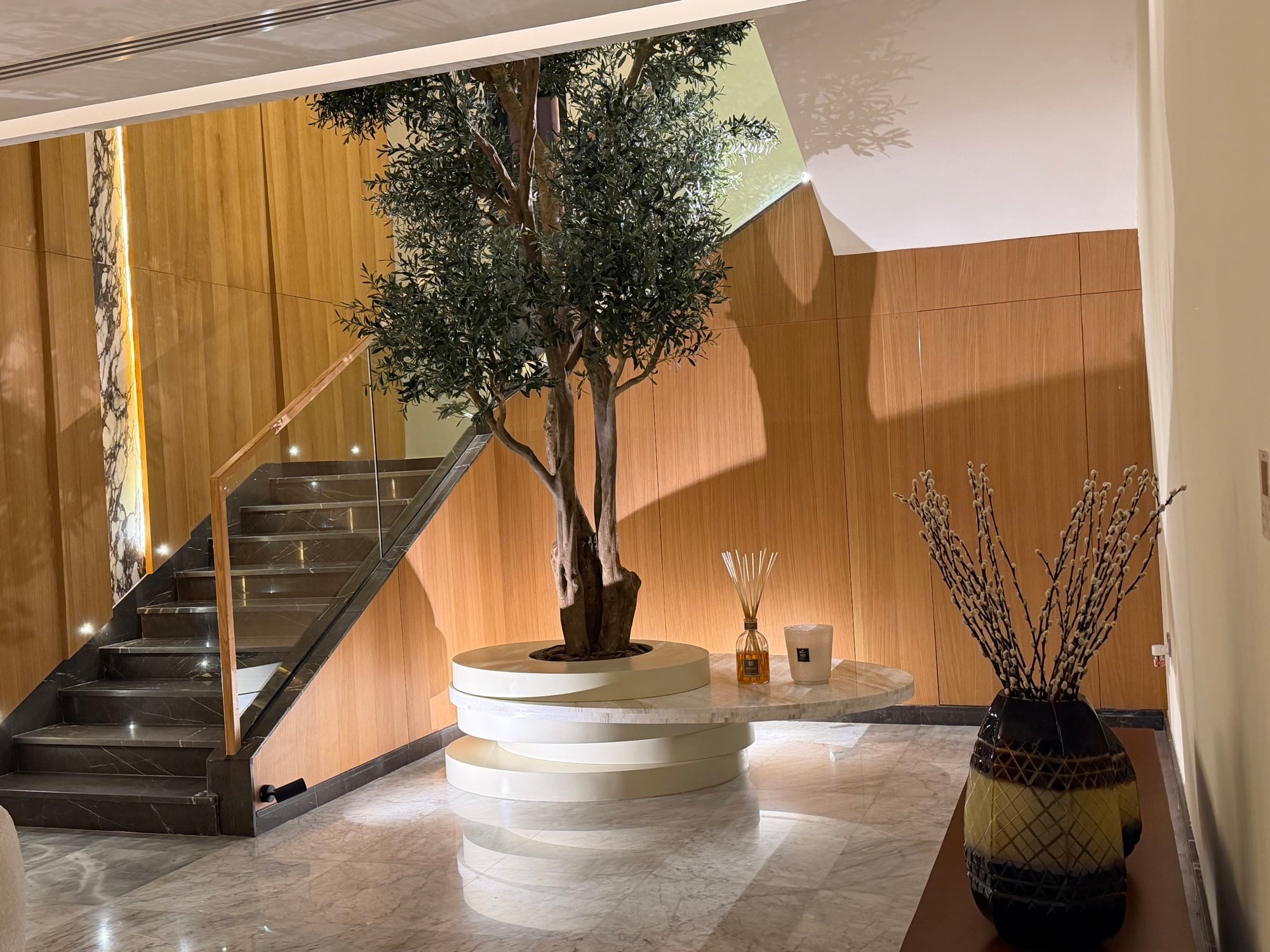 An olive tree on a stepped circular plinth beside a glass-balustraded staircase, against timber panelling, designed by Radwa Interiors.