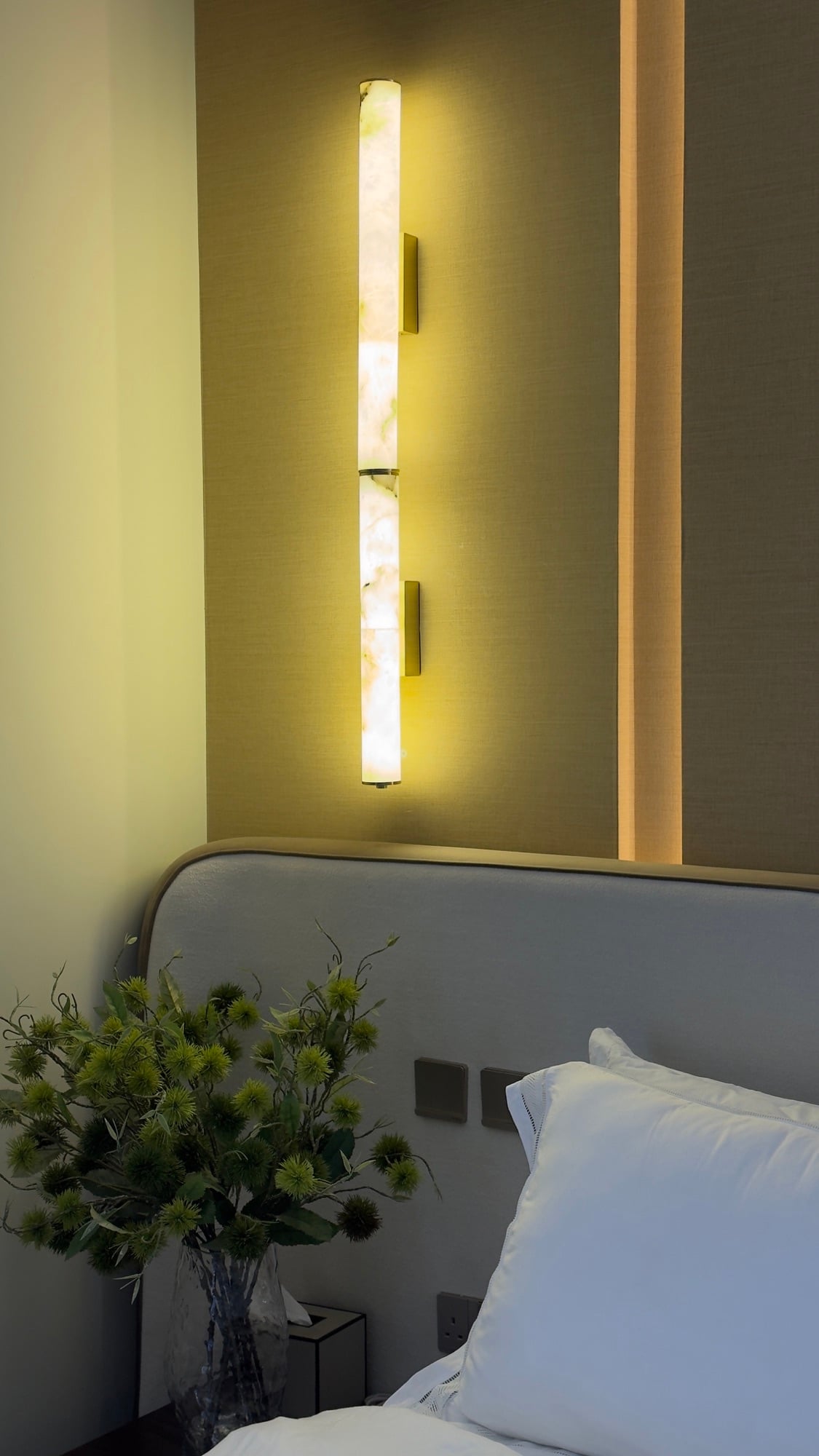 A backlit onyx wall light beside an upholstered headboard, with a vase of green thistles on the nightstand, designed by Radwa Interiors.