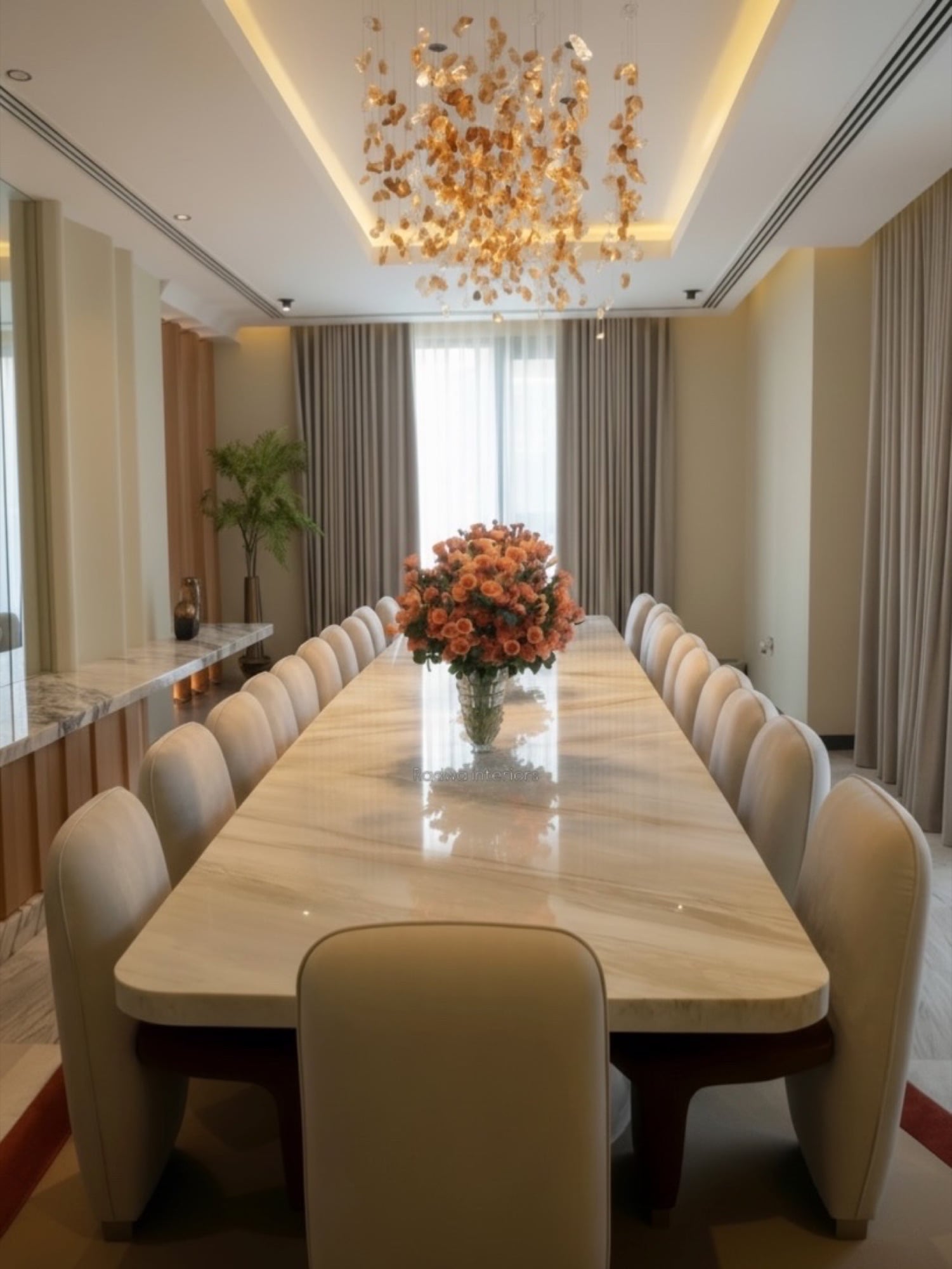 A long dining table under a chandelier of gilded leaves, lined with cream chairs and centred on a coral rose arrangement, designed by Radwa Interiors.