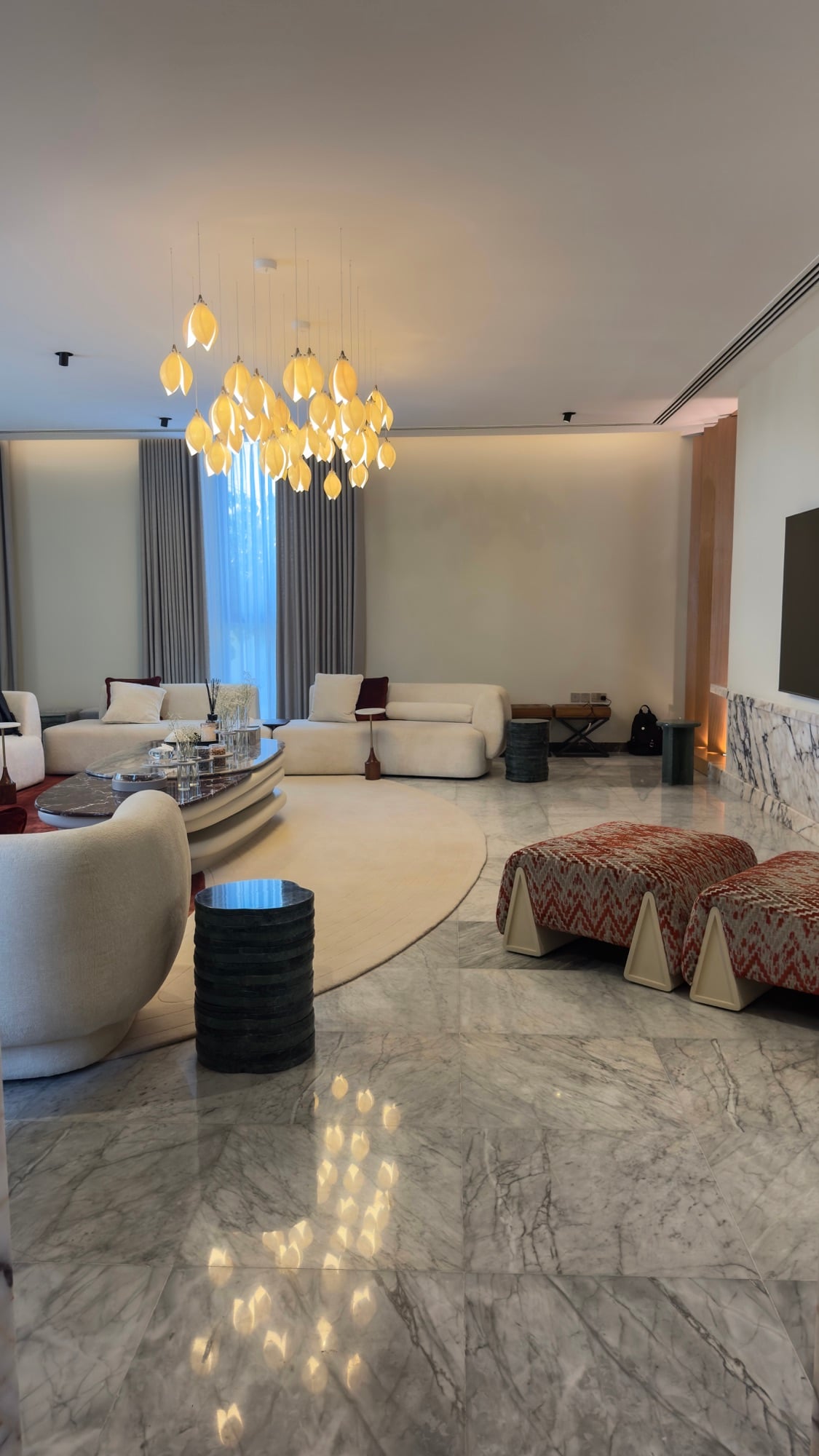 A living room reflected in polished marble flooring, with an amber leaf chandelier, cream seating and patterned ottomans, designed by Radwa Interiors.