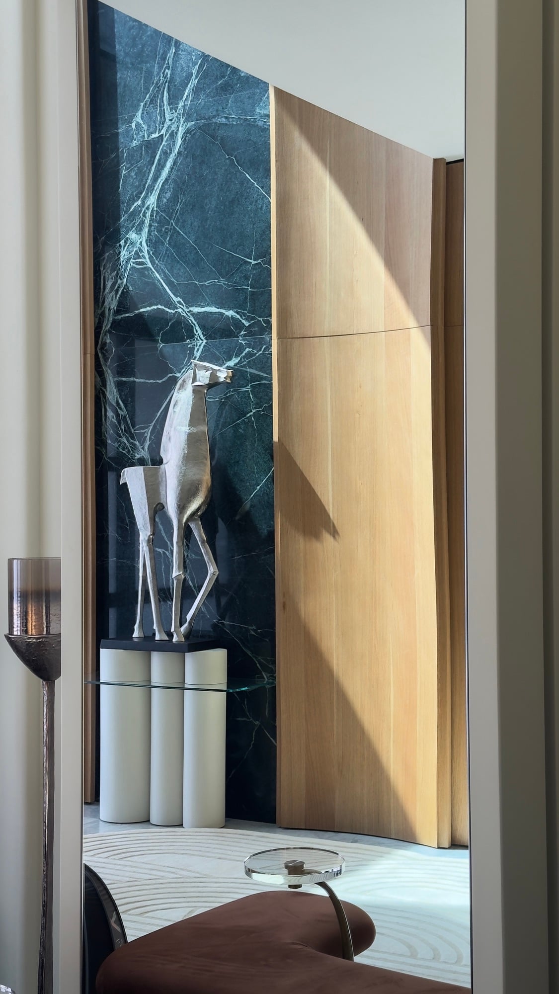 A silver deer sculpture on a fluted plinth against dark green marble, seen through a doorway past a leather bench, designed by Radwa Interiors.
