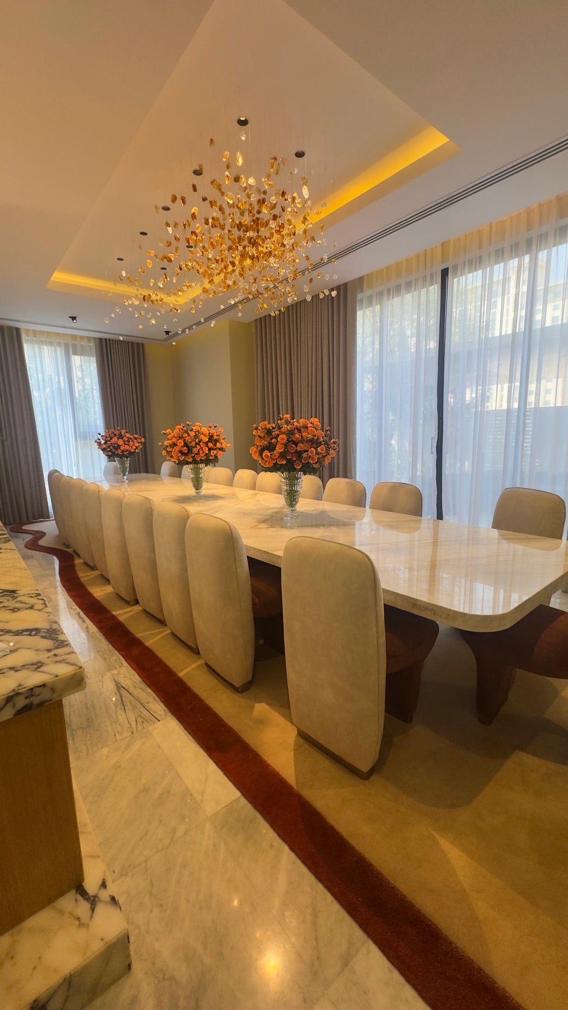 A dining table with cream high-backed chairs on a red-bordered rug, under a dense gilded chandelier, designed by Radwa Interiors.