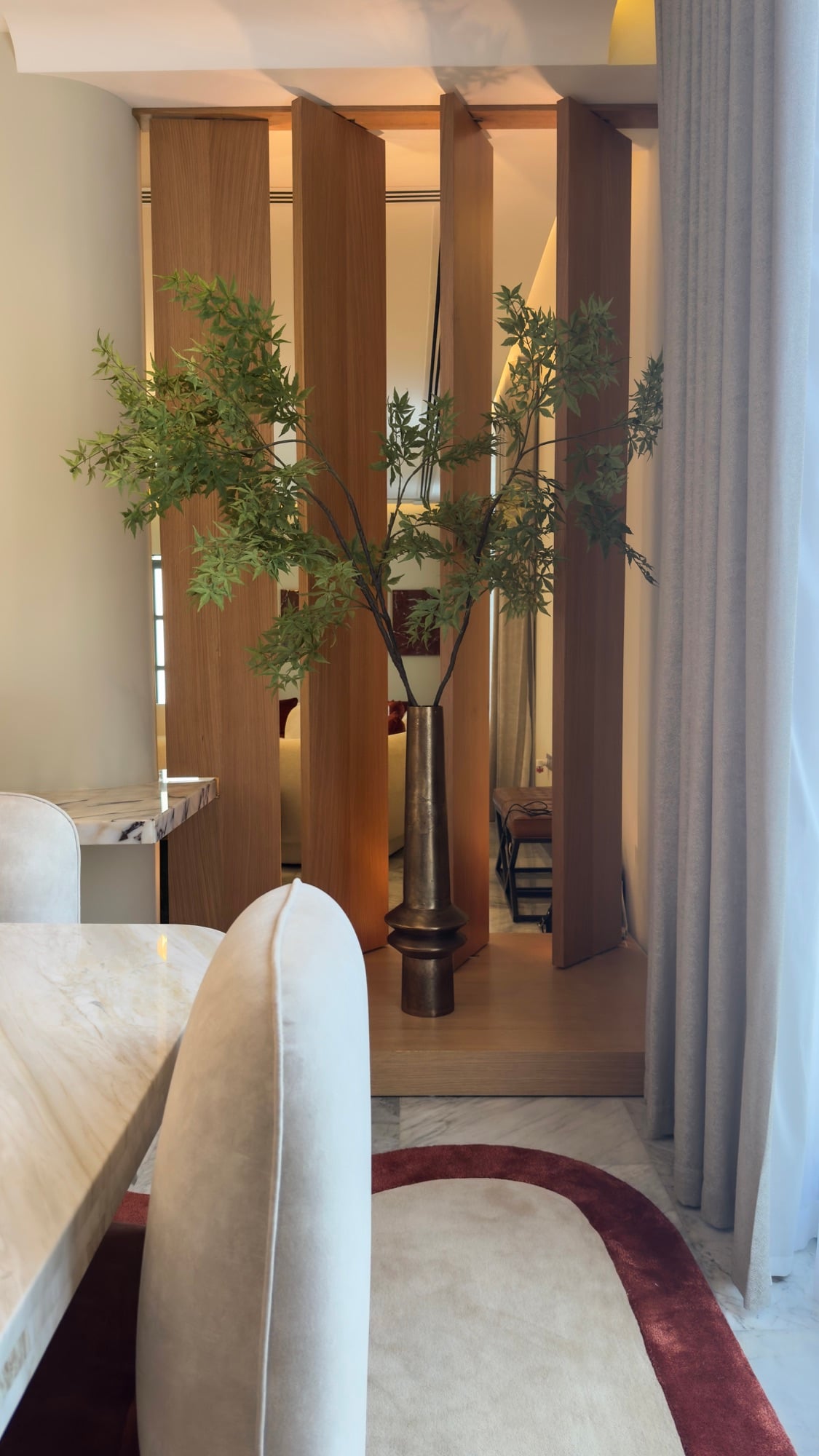 A slim tree in a bronze vessel between timber fins, seen past a stone dining table and cream chairs, designed by Radwa Interiors.