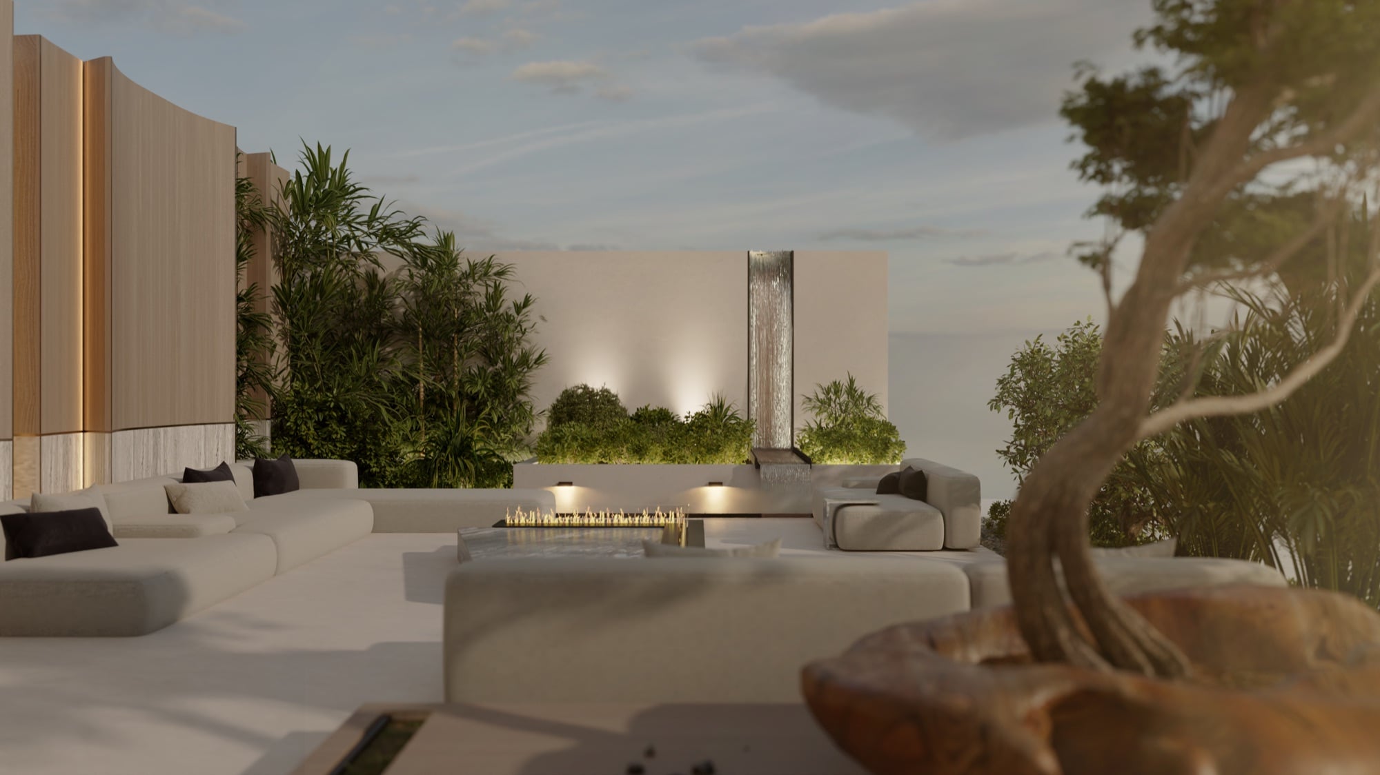 An evening terrace with cream outdoor seating around a fire table, beside a water blade and a sculptural tree, designed by Radwa Interiors.