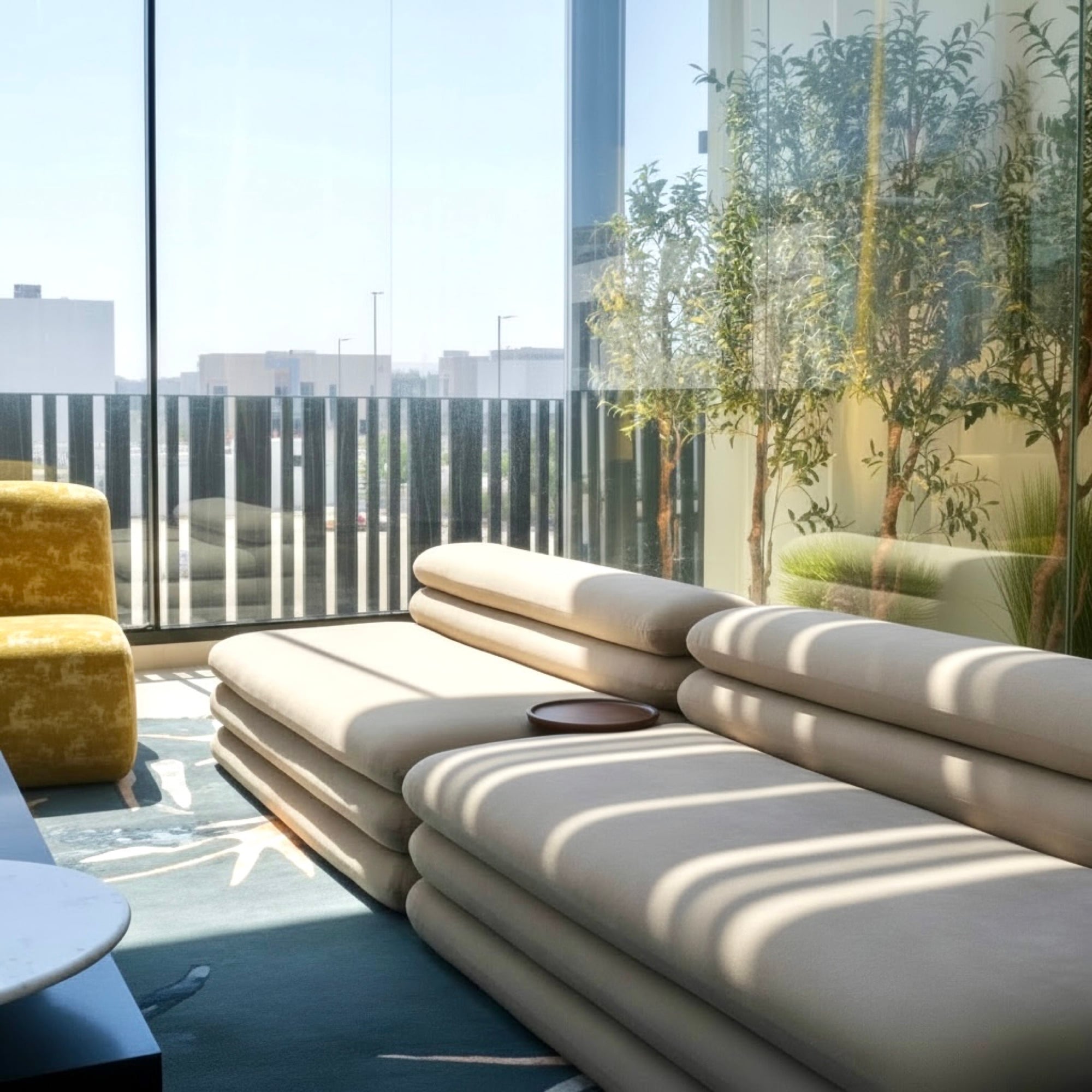A stacked cushion daybed in a glazed corner, sunlight striping across it, with olive trees behind the glass, designed by Radwa Interiors.